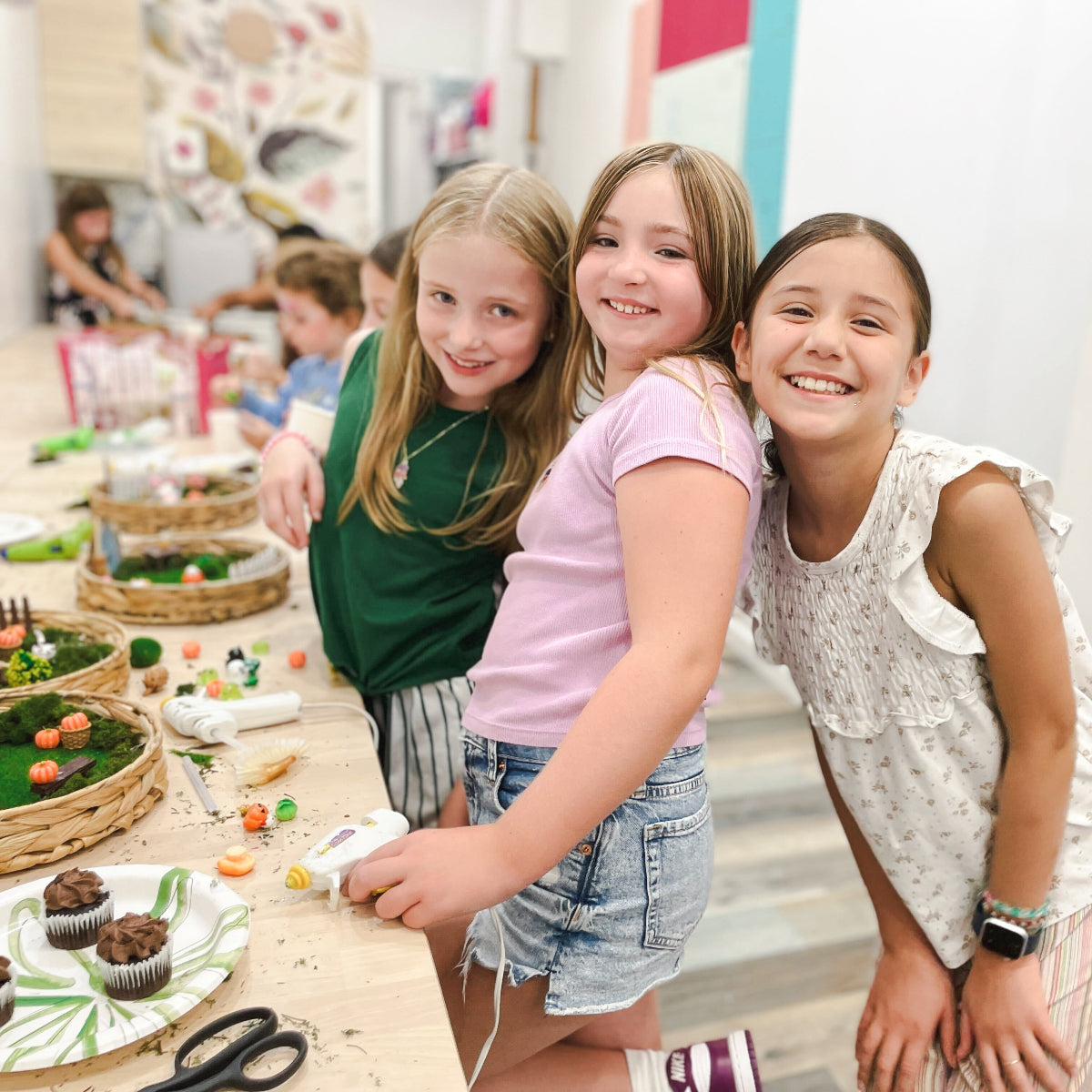 Three young girls standing around a table with food items in a casual setting.