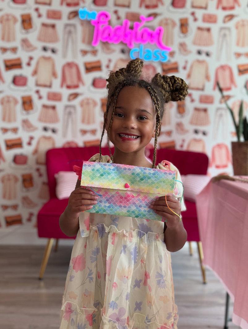 Young girl holding a colorful pouch in a room with a 'Fashion Class' sign on the wall.
