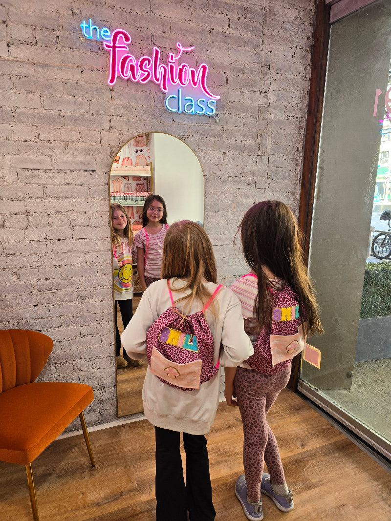 Two children standing in front of a mirror with a colorful 'The Fashion Class' sign.