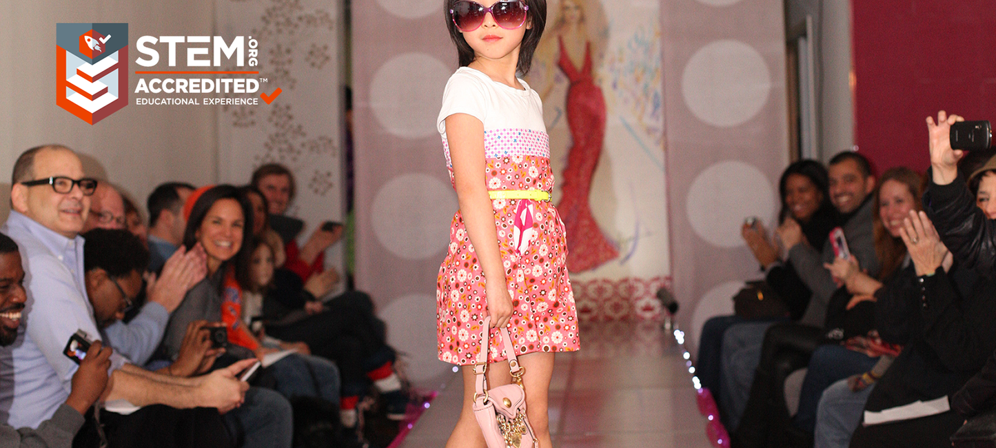 Young girl walking on a fashion show runway with 'STEM Accredited' logo in the corner.
