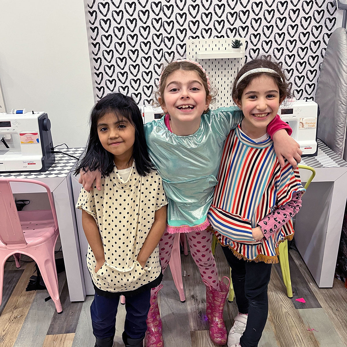 Three children posing together in a room with sewing machines and heart-patterned curtains.
