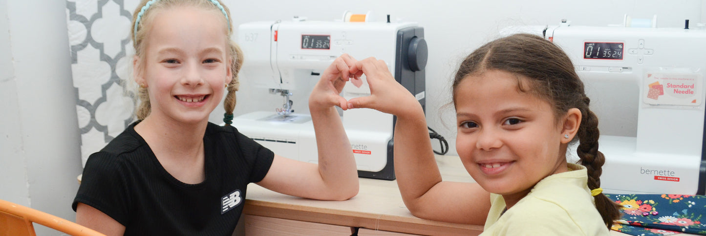 Two young girls making a heart in front of sewing machines.