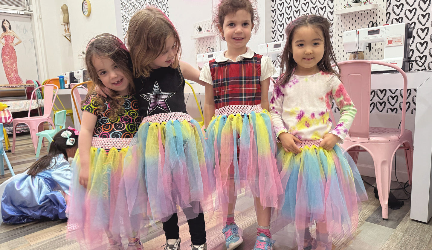 Toddler girls in tutus standing in front of sewing machines in class